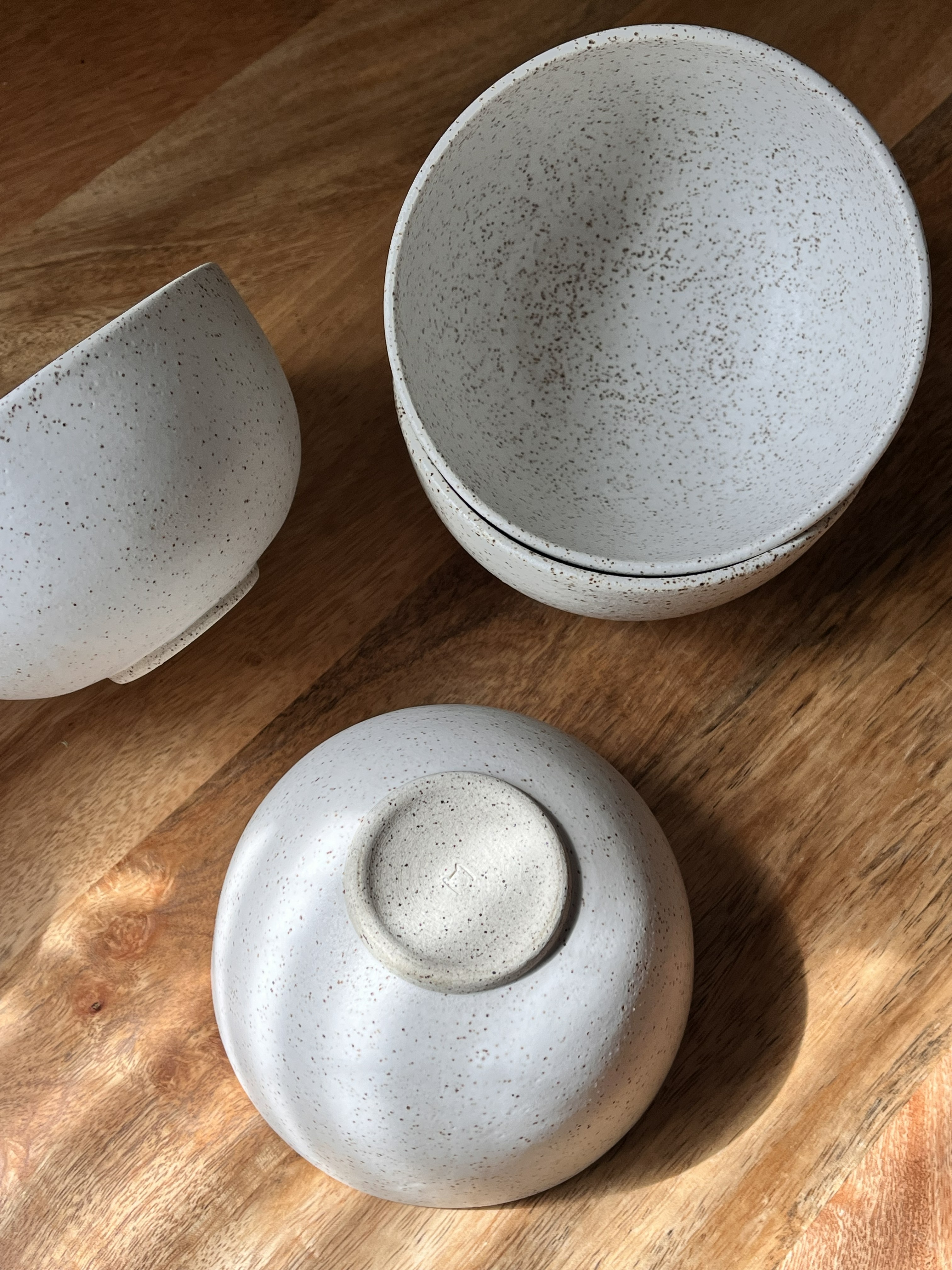 Speckled matte white handmade ceramic bowls on a warm wooden surface. One bowl is on its side, one is upside down to show the bottom of the bowl, and two are stacked on top of each other.