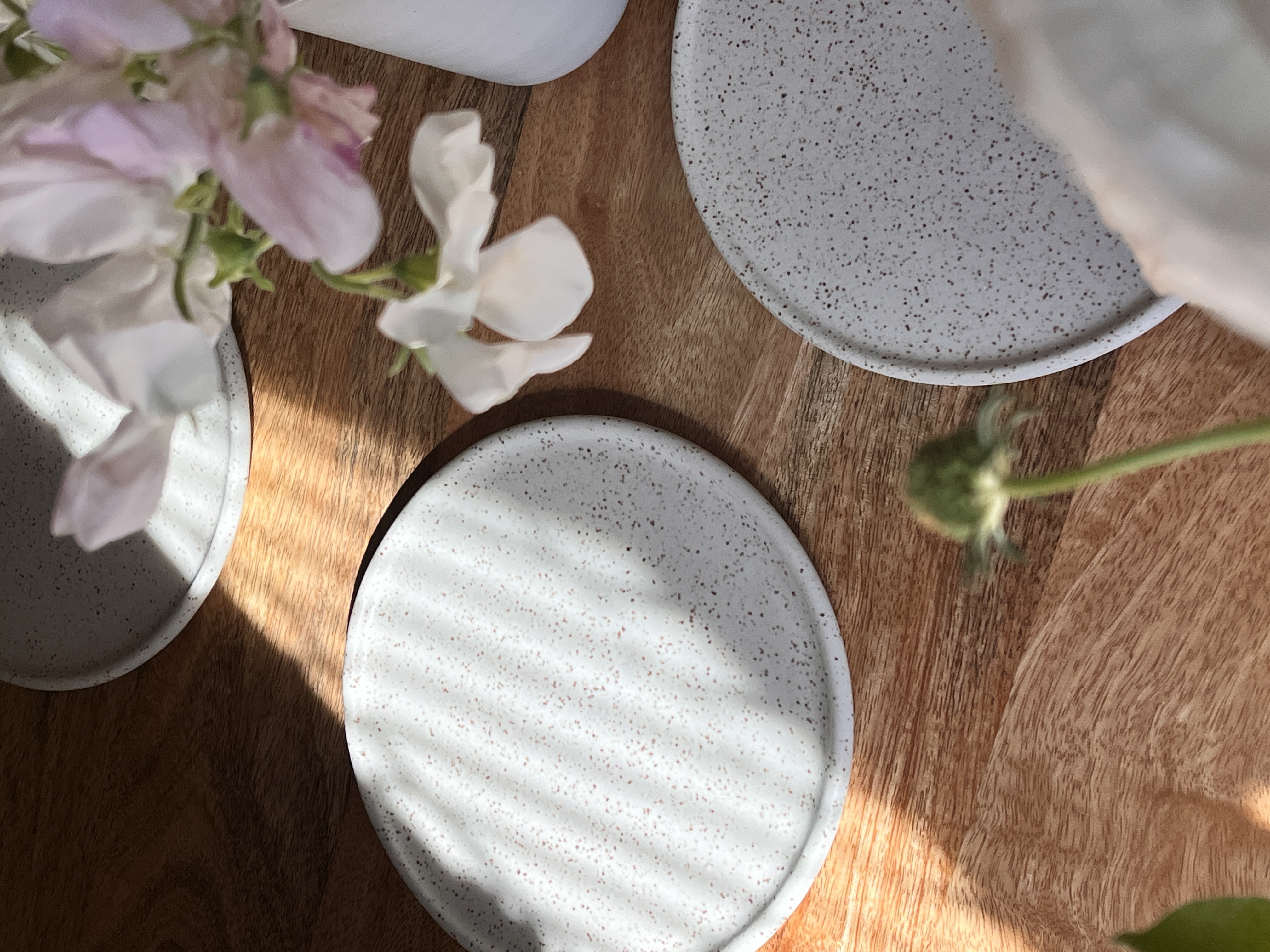 Mini speckled matte white handmade ceramic plates partially in the sunlight with pink sweetpea flowers in the corner.