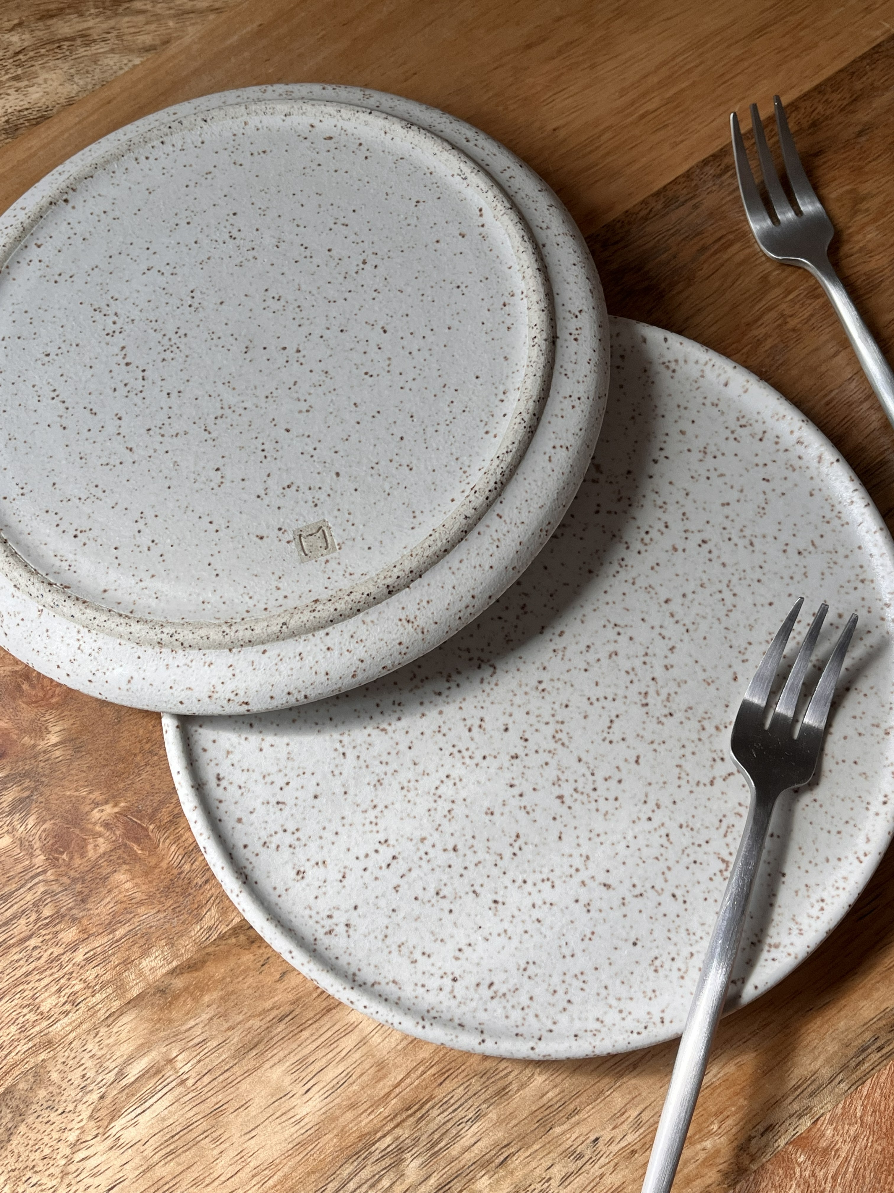 Close-up of two speckled matte white handmade ceramic dessert plates resting on top of each other. The top plate shows the bottom of the plate with a small signature stamp. Two silver mini forks rest on the right side of the plates.