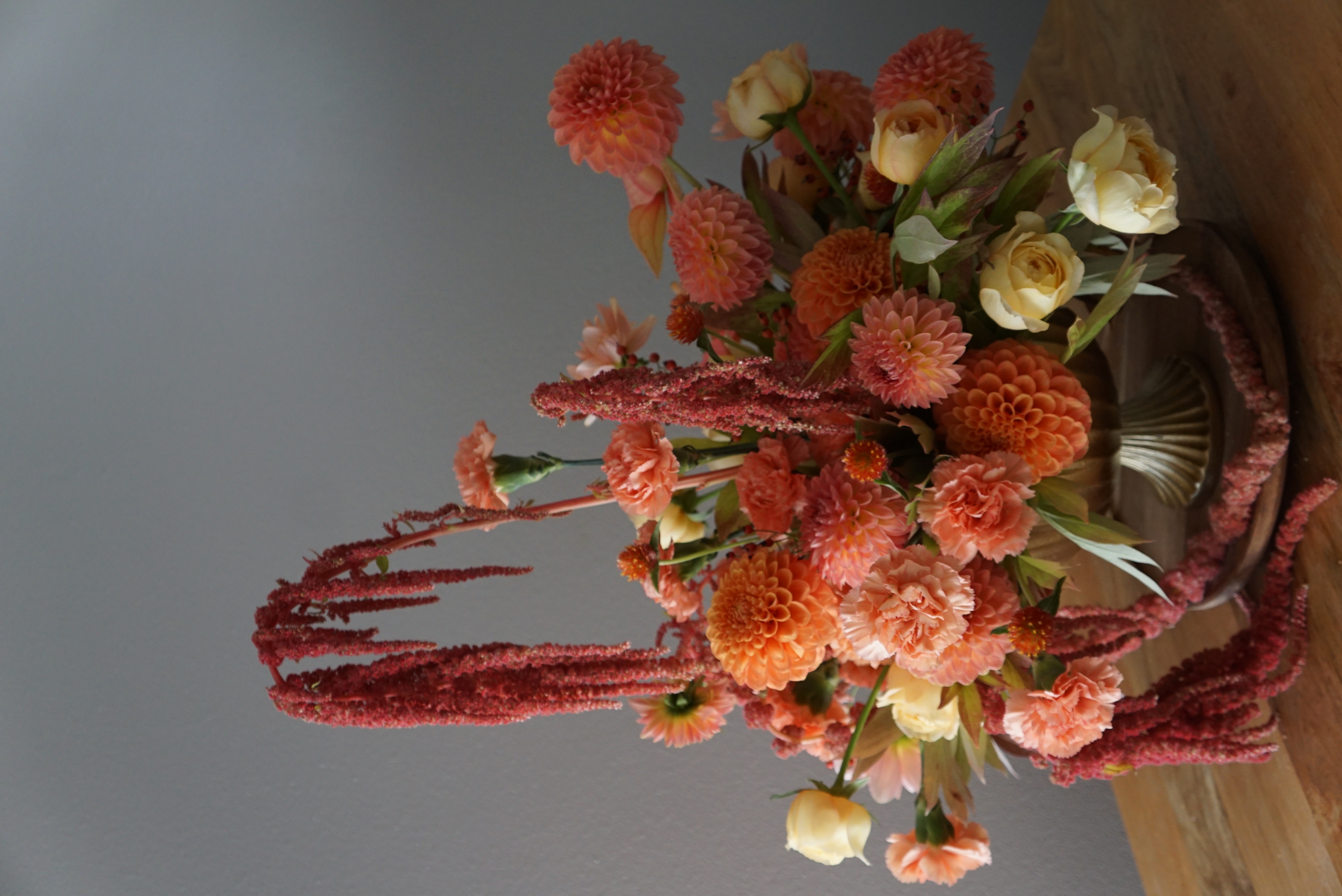 A bright fall arrangement featuring peachy carnations and dahlias, red rosehips and pincushions, pink and peachy anthuriums, orange-red gomphrenas, and caramel antike roses in a gold compote vase.
