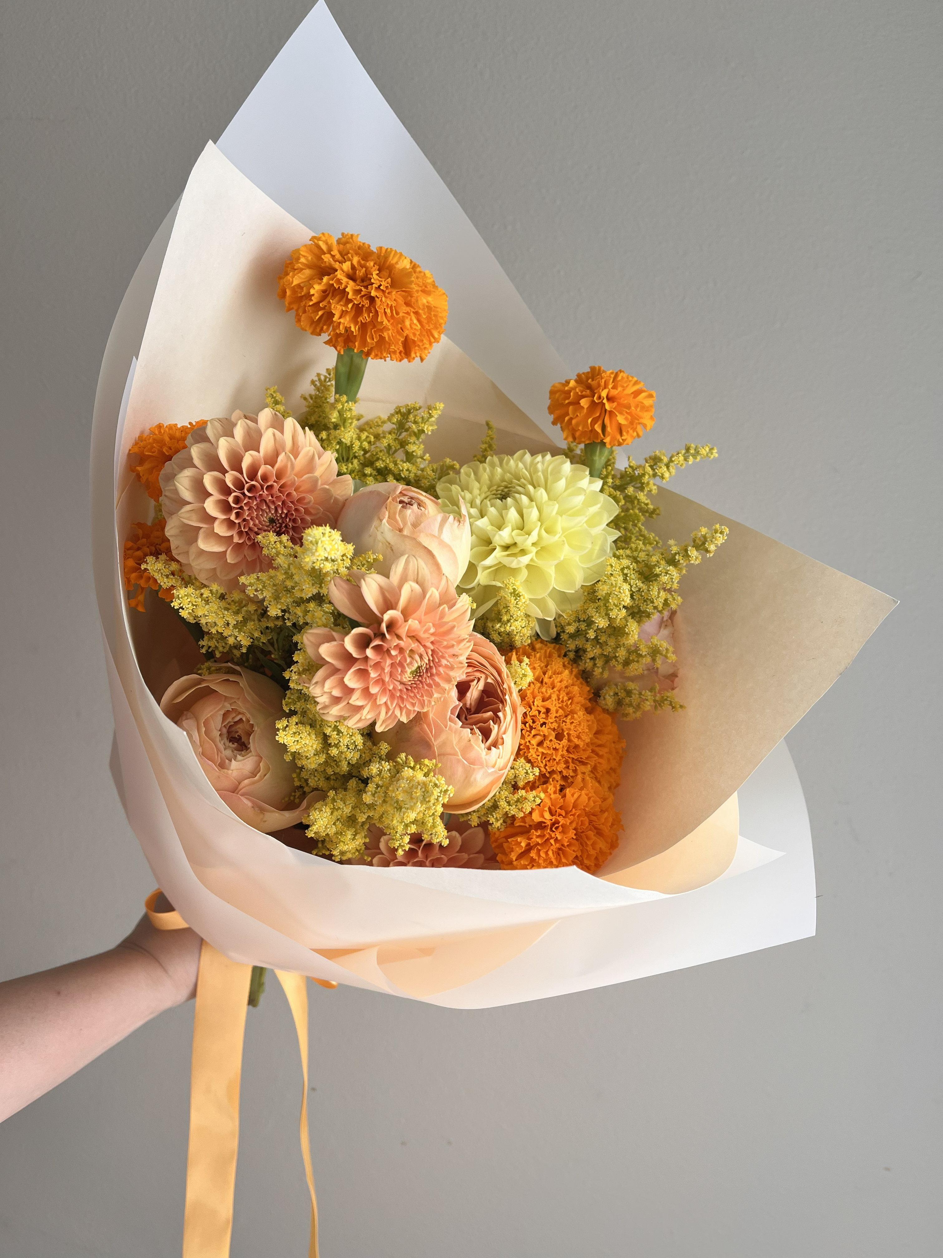 A hand-tied classic bouquet featuring orange marigolds, yellow and peach dahlias, yellow solidago, and peach roses wrapped in peachy and white wrapping paper and tied with a light orange ribbon.