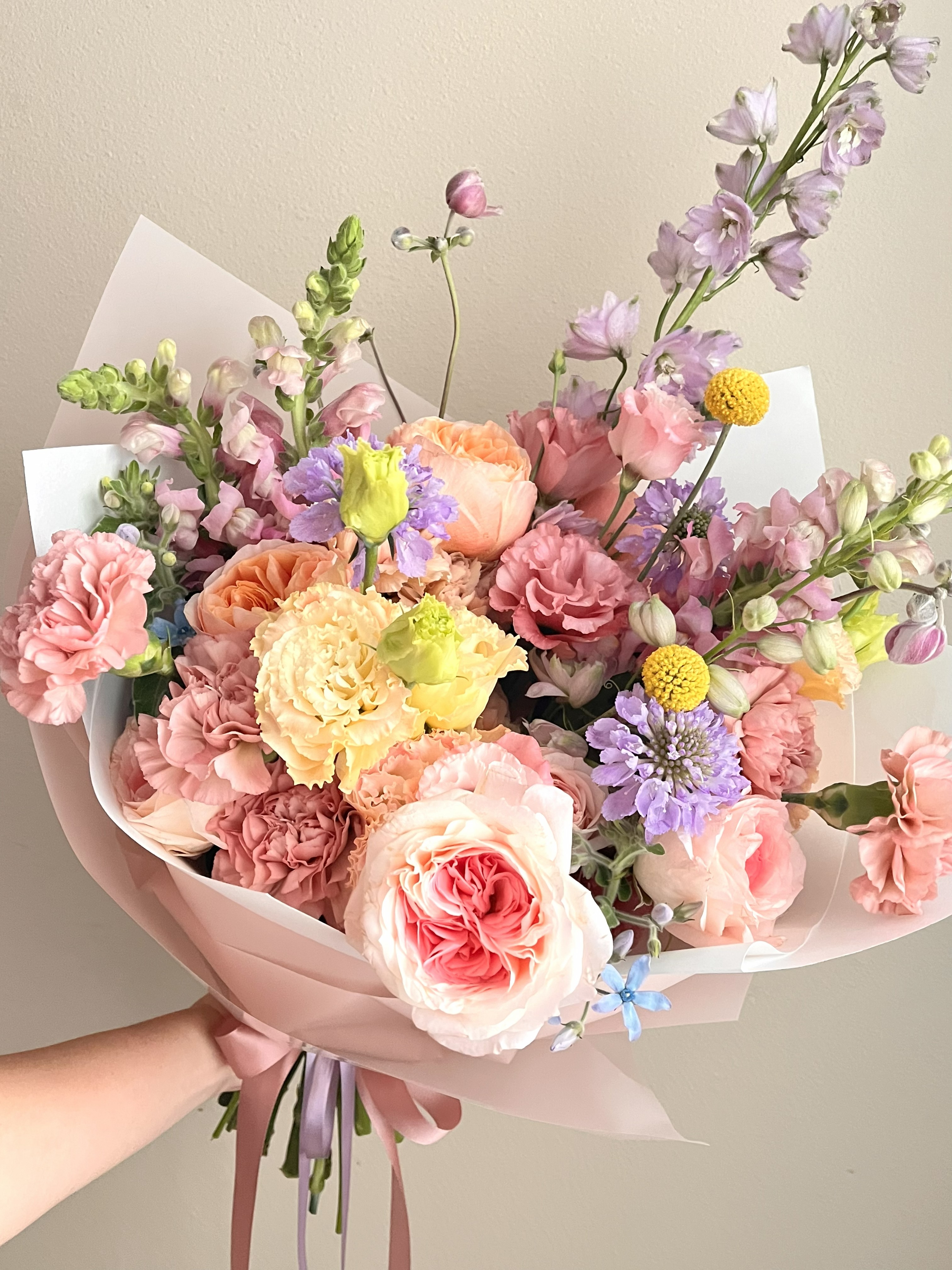 A colorful and pastel signature bouquet full of purple delphiniums and scabiosas, pink and peach roses, pink carnations, pink lisianthus, pink-purple snapdragons, yellow billy balls, and hints of blue tweeida. All wrapped in a pink and white wrapping with pink and purple ribbons.