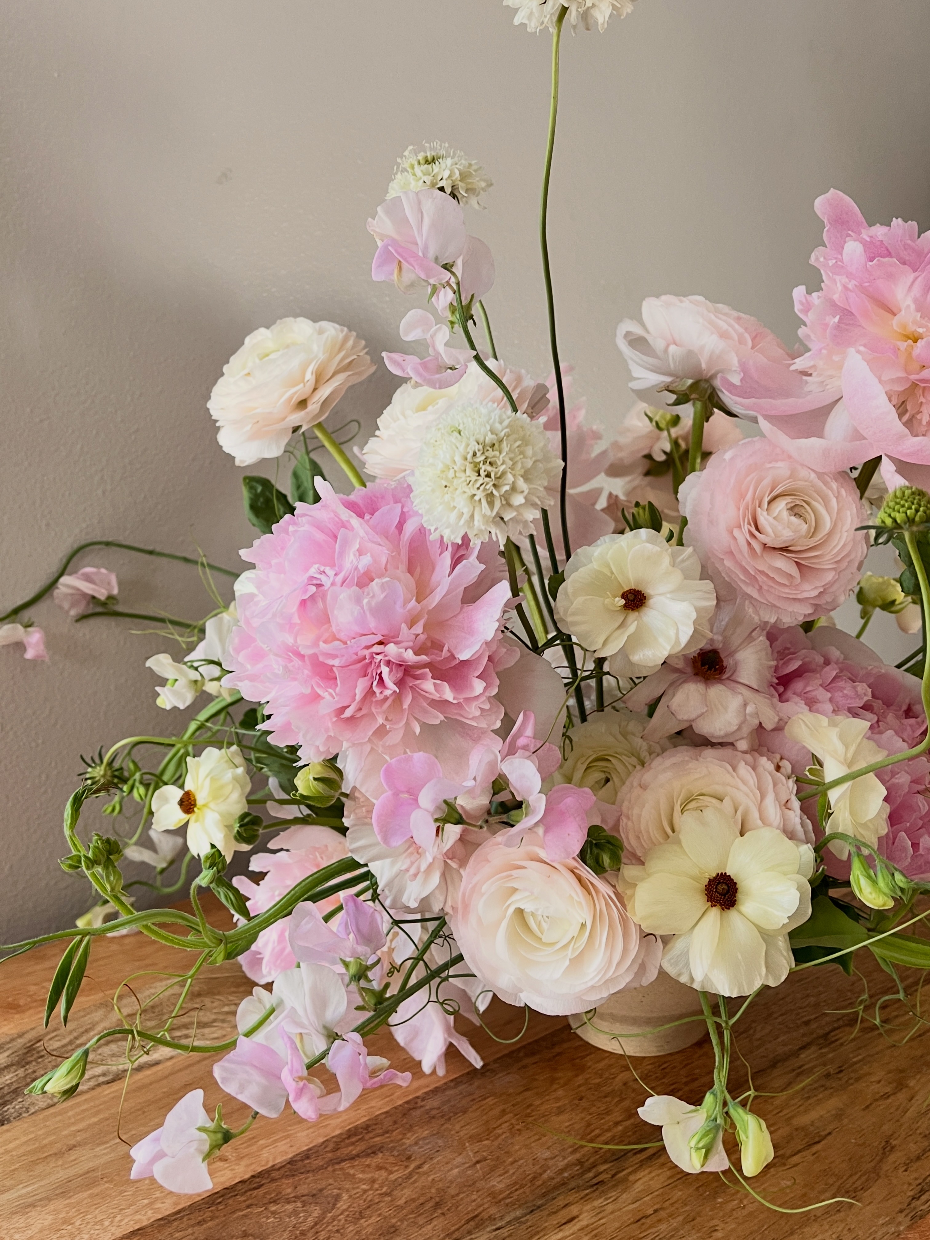 A signature flower arrangement with romantic lush garden vibes featuring pink peonies, pink and cream ranunculus, cream and yellow butterfly ranunculus, pink sweetpea vines, and white scabiosas.